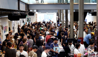 A busy indoor market or fair with crowded tables displaying various items. People are interacting, and a glass upper level shows a few more observers.