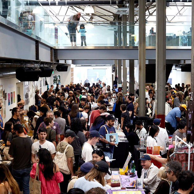 A busy indoor market or fair with crowded tables displaying various items. People are interacting, and a glass upper level shows a few more observers.