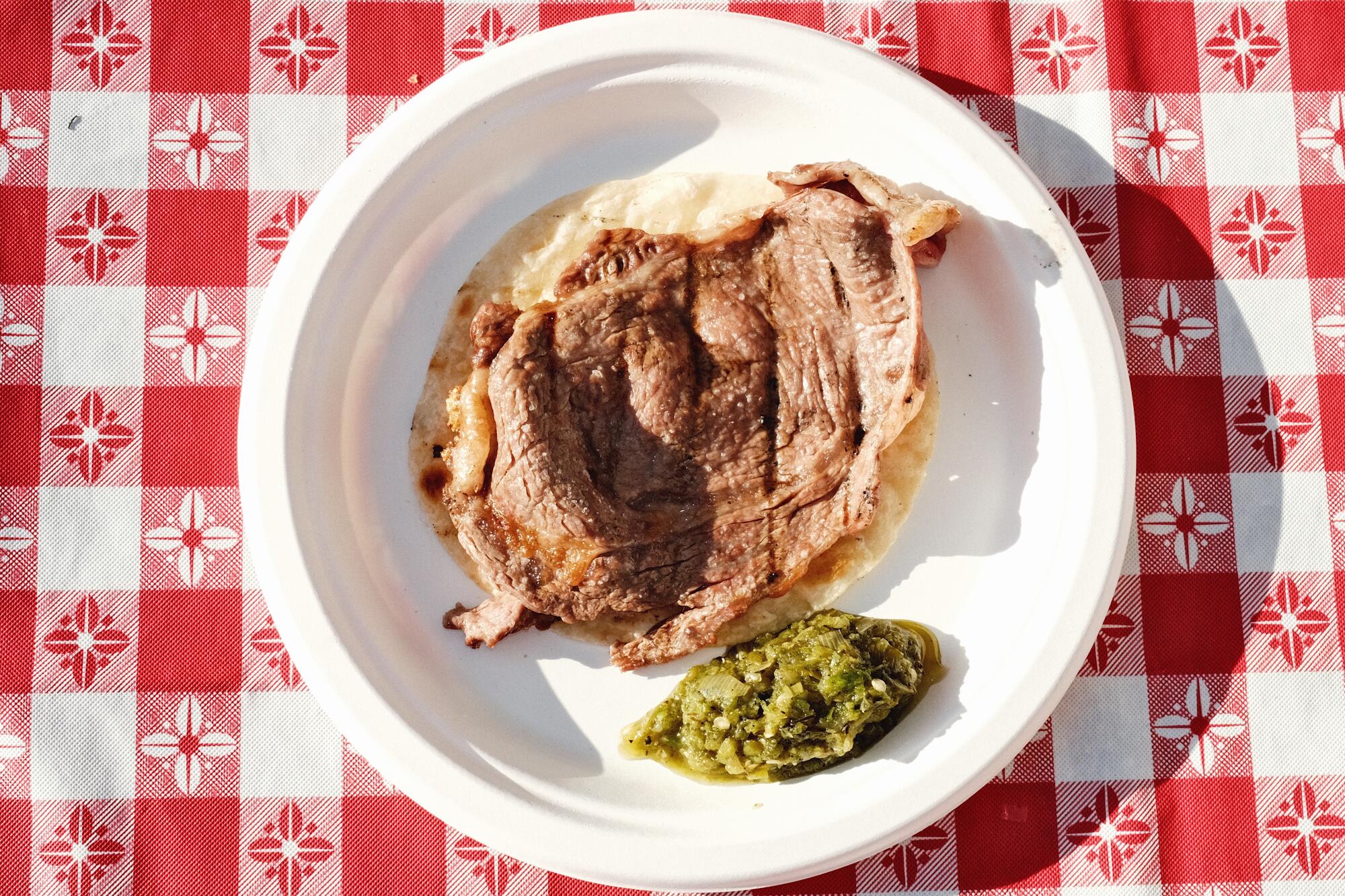 A rib-eye taco with a dollop of green salsa verse on a white paper plate atop a red-and-white checked table