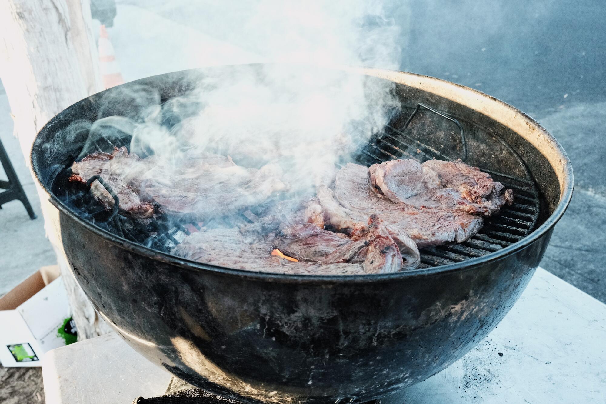 Plumes of smoke come from steaks on a black charcoal grill at Hollywood stand Asadero