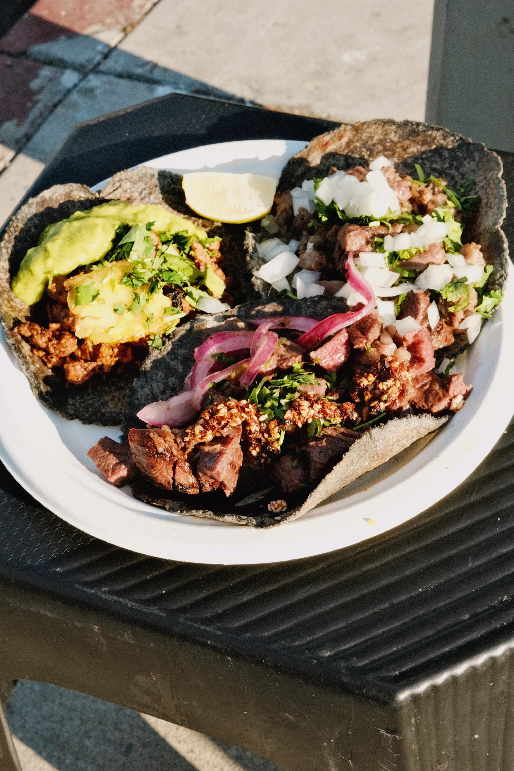 Three tacos in blue corn tortillas sit on a paper plate on a black plastic stool on the sidewalk