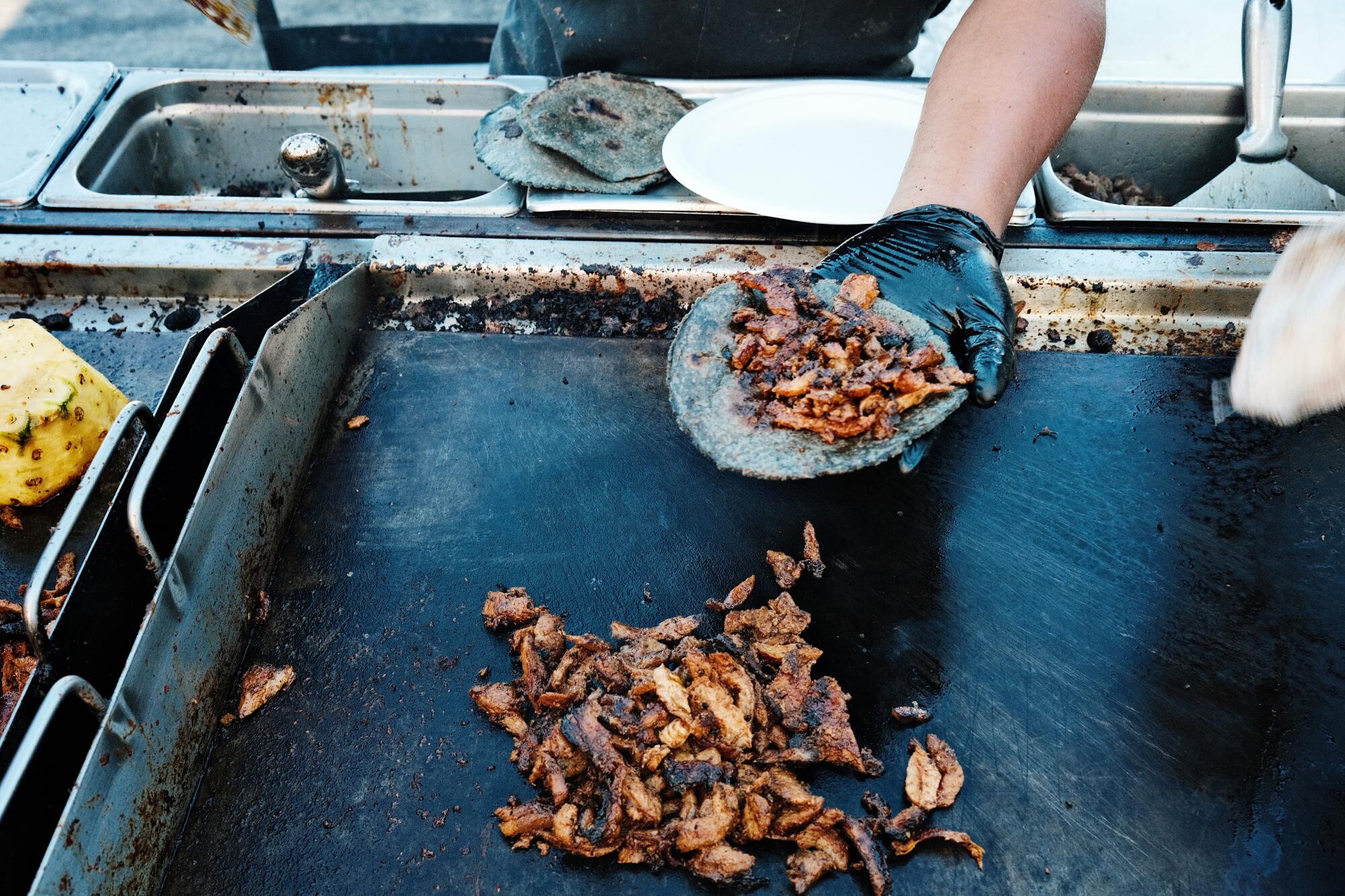A hand holds a taco over the flat-top grill where meat sizzles