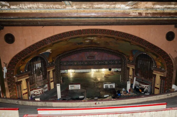 View from a theater balcony overlooking the stage of the historic Westlake Theatre. The proscenium arch is richly decorated with painted murals, ornamental trim, and arched side openings. The stage appears vacant and under renovation, with a dark floor, scattered equipment, and temporary structures visible. The auditorium shows signs of age, with worn finishes and peeling paint on the ceiling and walls, while ambient lighting illuminates the expansive interior.