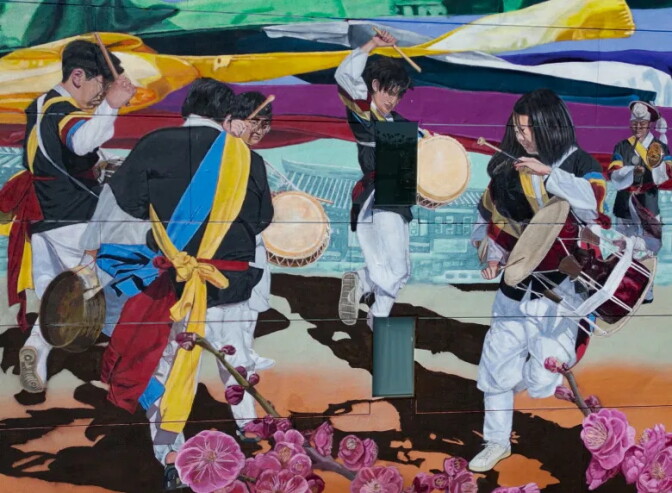 A close-up of the mural showing five Korean drummers in traditional white and black dress with yellow, blue and red sashes, playing hand drums and gongs. One younger drummer leaps mid-air in white sneakers. Faint outlines of traditional Korean architecture appear behind them, with pink plum blossoms blooming across the bottom.