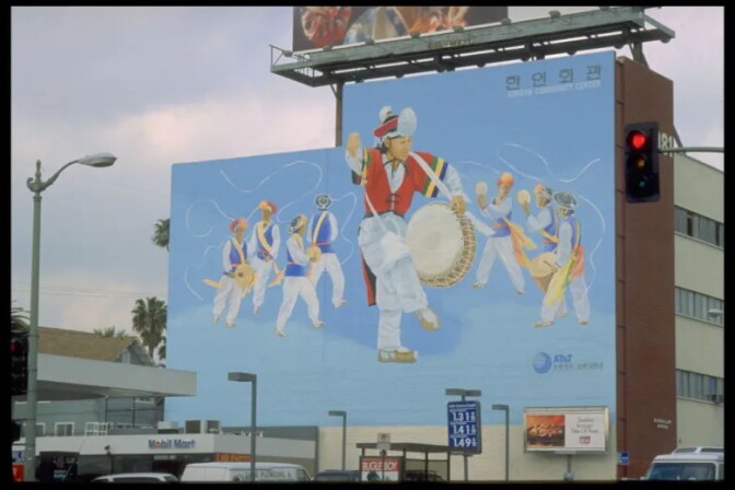 A large mural on the side of a building shows Korean drummers and dancers in white traditional dress with red and blue vests performing against a pale blue sky. A central figure leaps mid-dance holding a barrel drum, ribbons streaming from a hat. Korean text and "Korean Community Center" appear at the top right. Below, at street level, are a Mobil gas station, a traffic light and a billboard.
