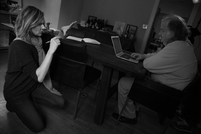 A female presenting person kneels in front of a male presenting person who sits at a desk in front of a computer.