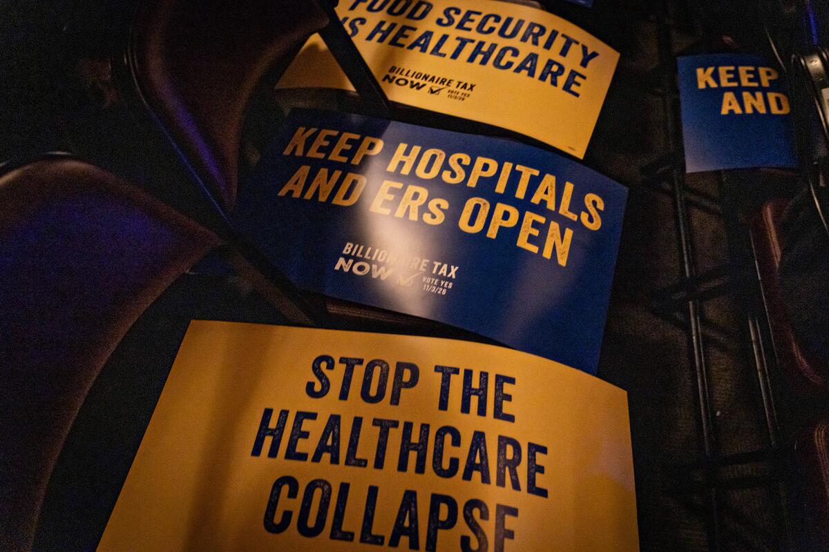 Billionaire Tax Now signs sit atop folding chairs. One says "Keep hospitals and ERs open."