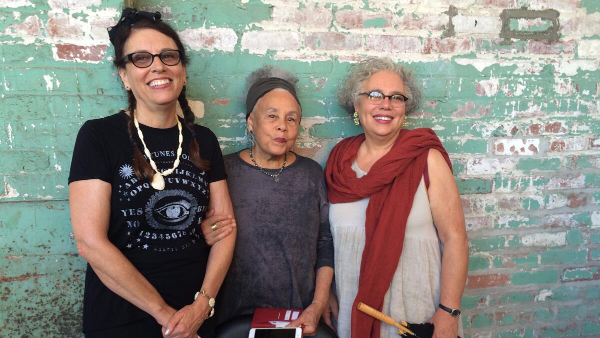 Betye Saar, flanked by her daughters, stands before a brick wall with fading paint.