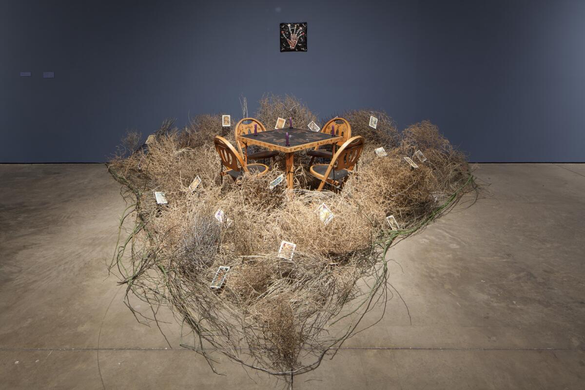 A museum installation shows tumbleweeds surrounding a table for four in front of a wall painted blue.