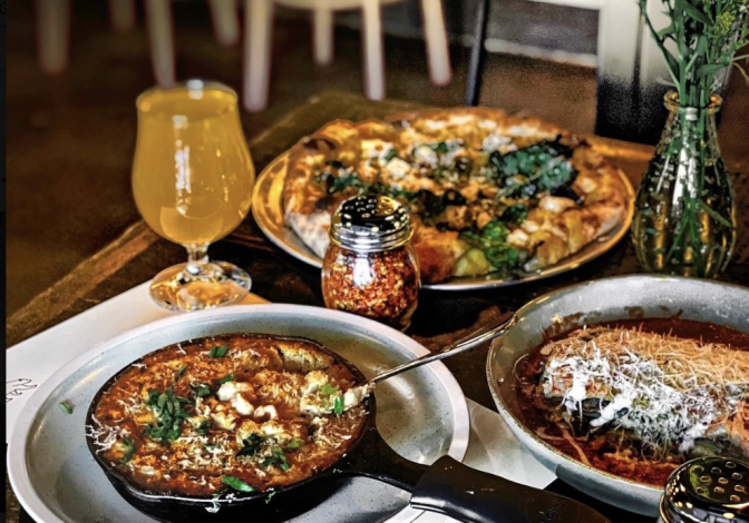 A selection of dishes on a wooden table include a cast iron skillet full of mac and cheese, a metal dish with pizza on top and another metal dish containing lasagna. There's also a glass of amber looking kombucha.