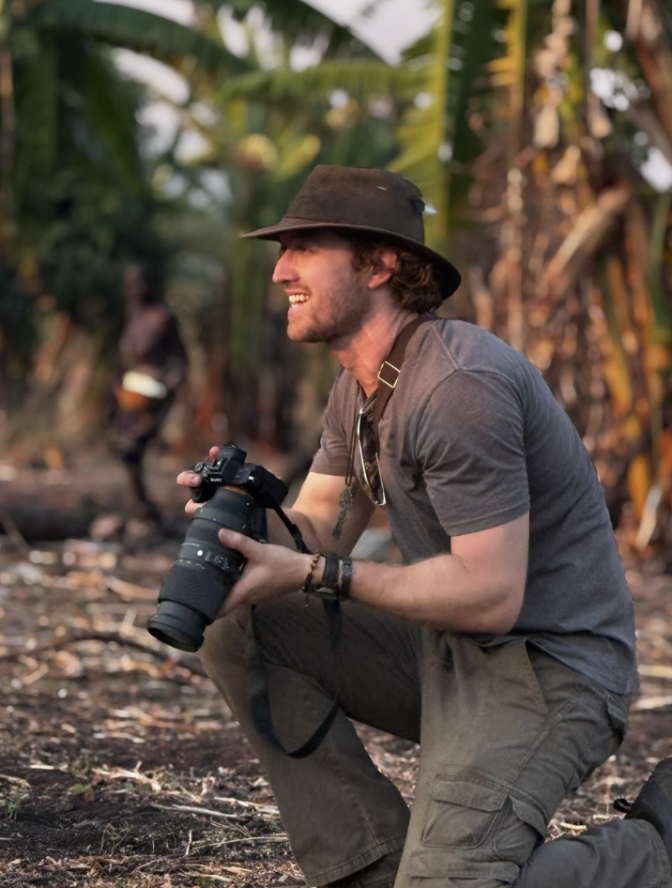 A light skinned man is kneeling down in a forest. He's in profile view and is holding a camera.
