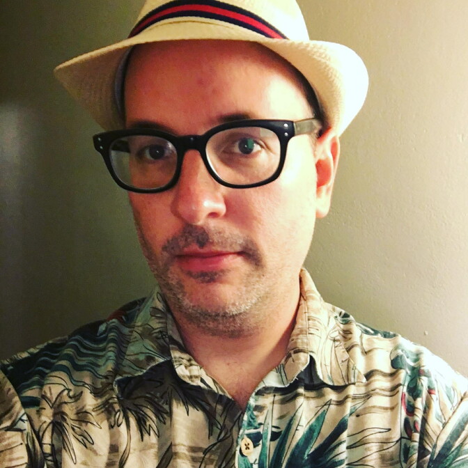 A light skinned man wearing black glasses and a sunhat looks at the camera.