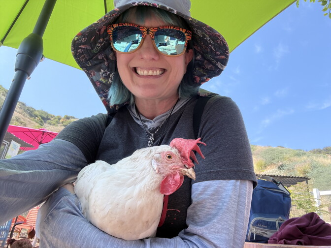 A light skinned woman wearing sunglasses and a sunhat is smiling at the camera. She's holding a white chicken.