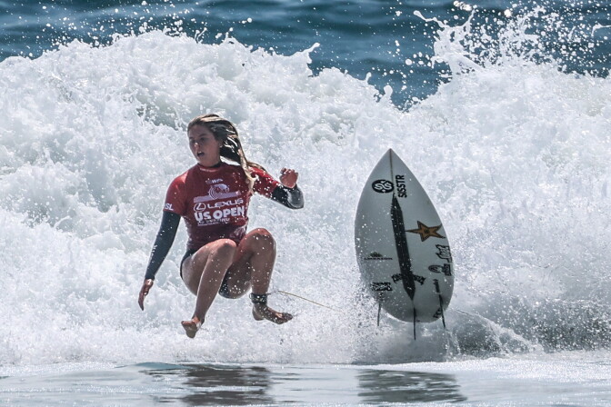 A light-skinned woman wearing a red surfing suit hovers above a crashing wave while her surfboard flies next to her. 