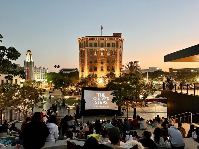 A large brick building lit up behind a plaza. In front of it is a large projector screen that reads "The Culver Steps." People sit on the steps and watch. 