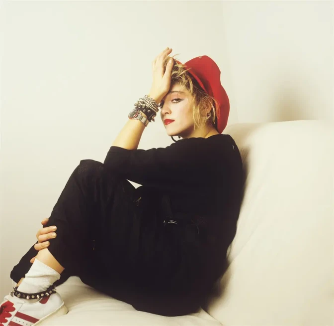 A light-skinned woman in a red hat and black clothes sits on a white couch with her hand on her forehead. 