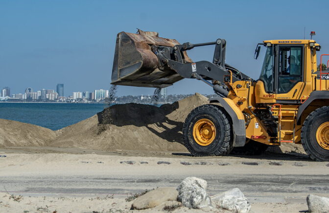 A tractor dumps a pile of sand on a beach. Tall buildings are in the distance separated by a body of water.