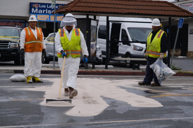 Crews wearing safety vests apply dirt on a street with oil on the pavement. A small plaza and bust stop are behind them.