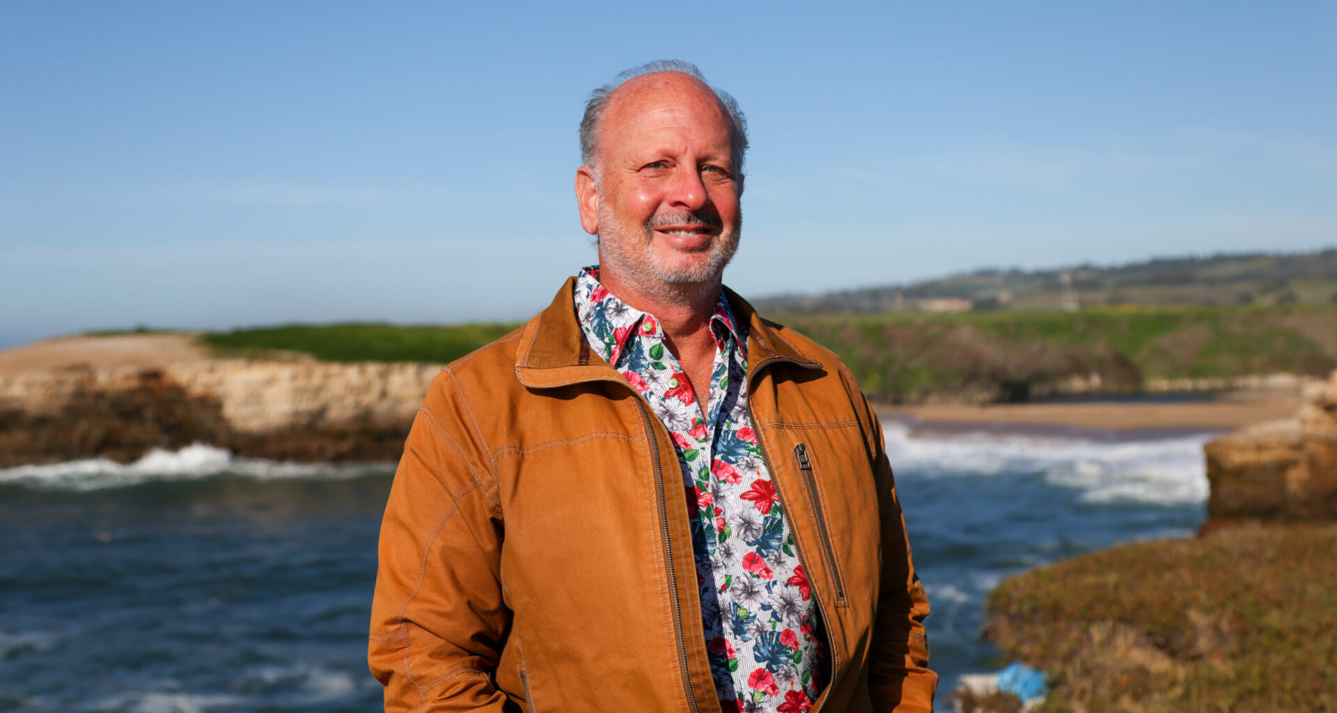 Mike Beck in a floral shirt and rust jacket by the Monterey Bay coastline.