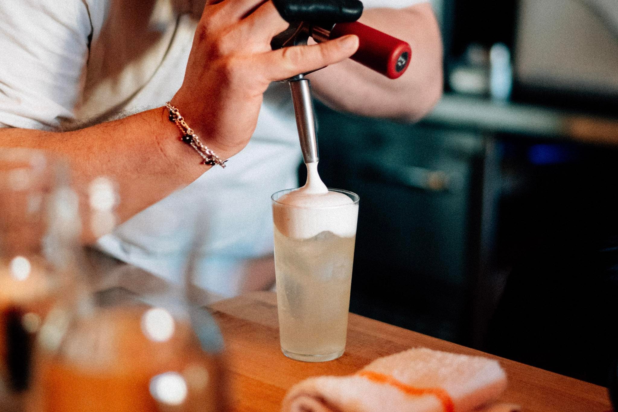 Person making cocktail in highball glass at Lynx, topping it with foam.