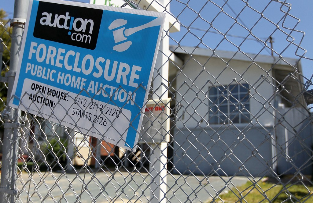 A foreclosure sign for a public home auction hangs on a chain-link fence in front of a house.