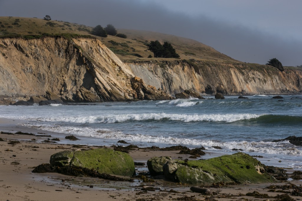 Waves break on a sandy beach with two moss-covered boulders in the foreground, and steep cliffs of varying colors in the background.