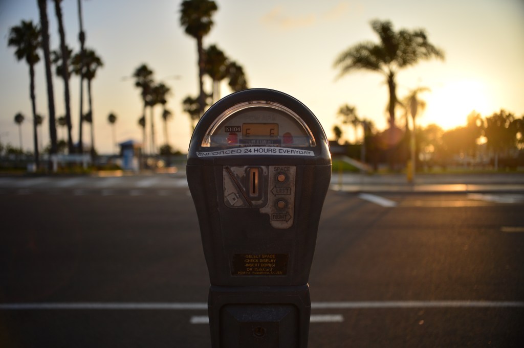 A parking meter with the text "ENFORCED 24 HOURS EVERYDAY" on its face, against a blurry sunset backdrop with palm trees.
