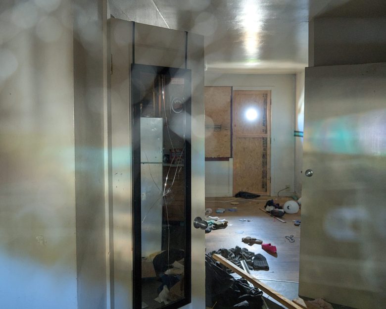 A cluttered room with scattered clothes and debris on the floor, a boarded-up window, and light shining through a round hole in the boarded area.