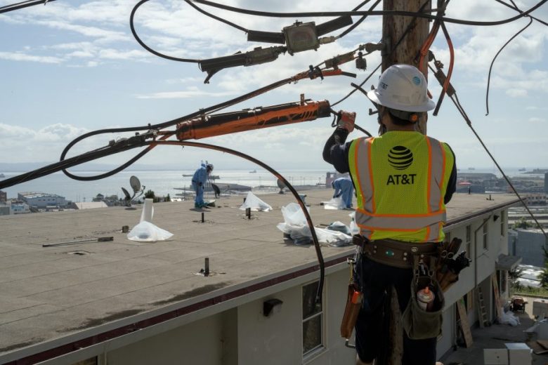A utility worker wearing an AT&T vest and helmet repairs wires on a pole, while two people work on a nearby rooftop with a view of water and ships in the background.