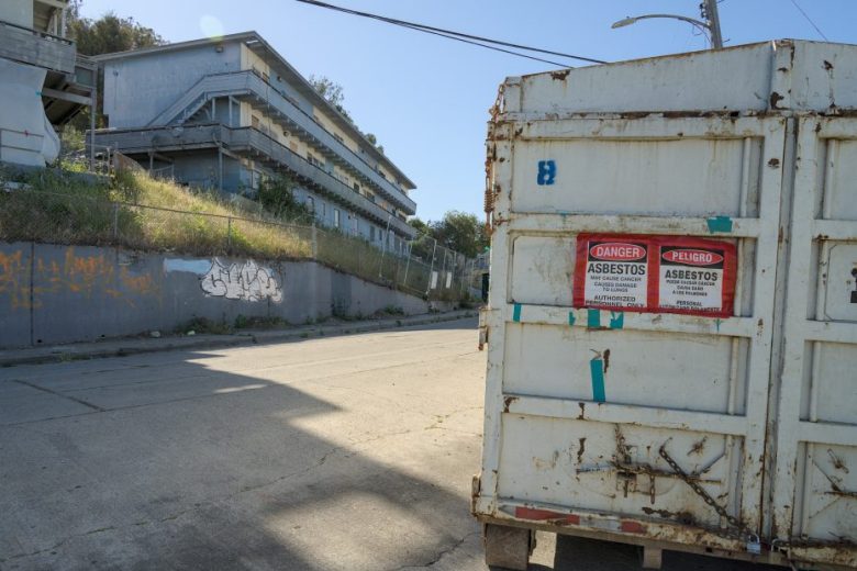 A metal dumpster with asbestos warning signs is on a deserted street near a multi-story building and a graffiti-covered wall.