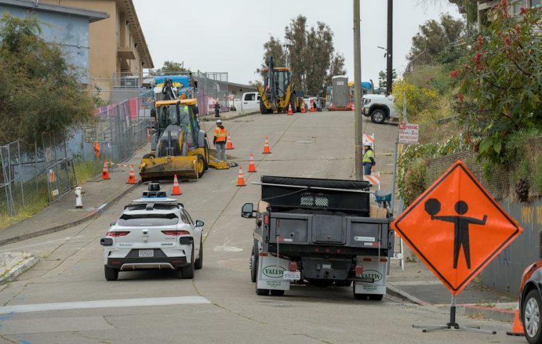 Road construction scene with cones, workers, two excavators, a white SUV, a black utility truck, and an orange "men at work" sign on a sloped street near buildings and trees.