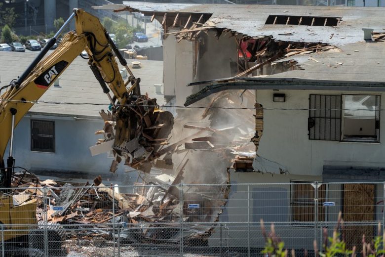 A yellow excavator demolishes part of a two-story building, with debris and dust falling to the ground.