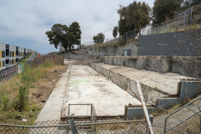 Abandoned empty swimming pool with cracked concrete, overgrown grass, and a chain-link fence in the foreground; buildings and trees in the background.