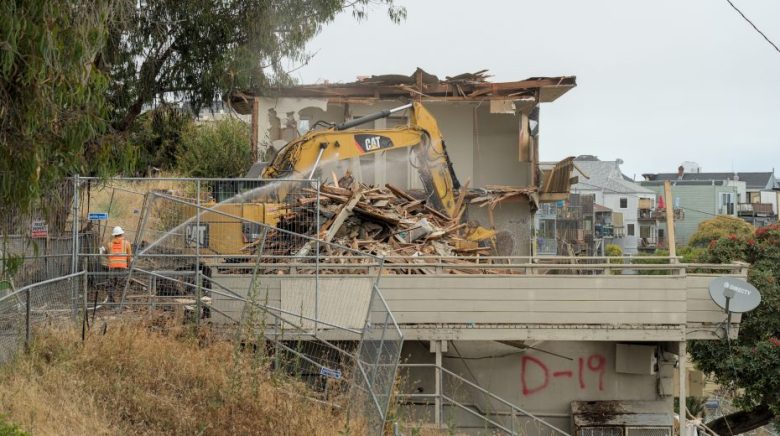 A yellow excavator demolishes a two-story house as a worker sprays water to control dust. Debris and construction fencing are visible, with "D-19" painted on a wall in red.