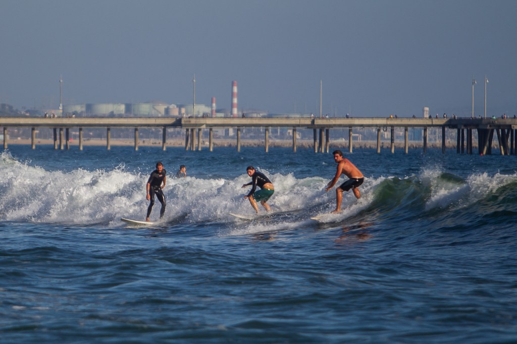 Three surfers ride waves at Venice Beach with a pier in the background.