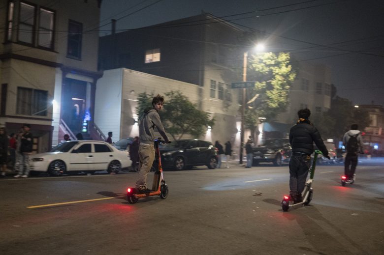 Three people ride electric scooters on a city street at night, with parked cars and buildings visible in the background.