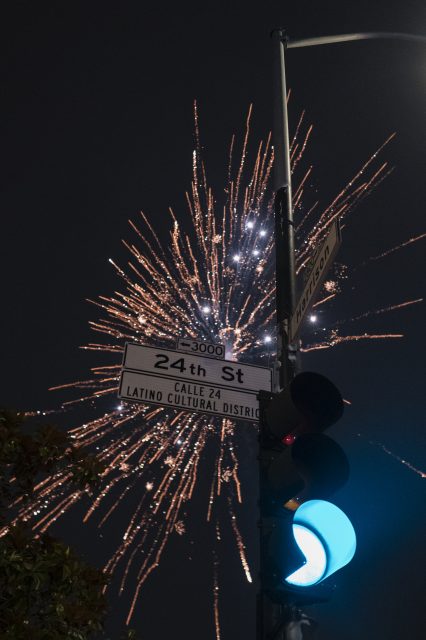 Fireworks explode in the night sky behind street signs for 24th St and Harrison St in the Latino Cultural District, next to a green traffic light.