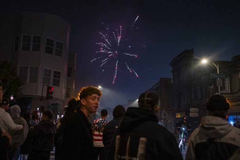 A group of people stand on a city street at night, watching fireworks exploding in the sky above buildings and street signs.