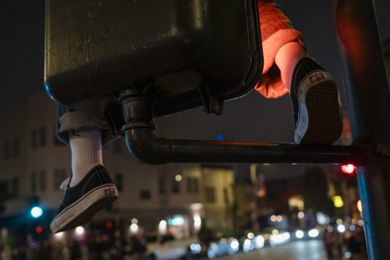 A person wearing black sneakers and white socks sits on a traffic signal box above a street at night, with blurred city lights and buildings in the background.