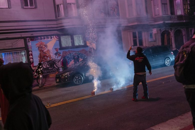 A person stands in the street at night taking a photo or video of fireworks or smoke devices, with murals and parked cars visible in the background.