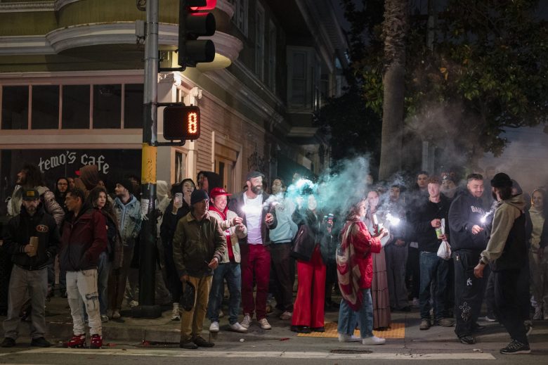 A group of people stand on a street corner at night, some holding drinks and cigarettes, with smoke visible in the air and a red traffic signal in the background.