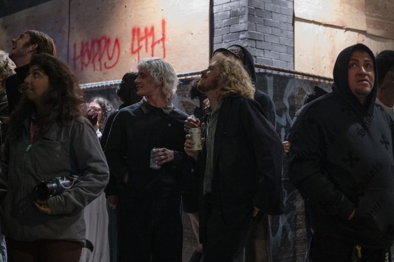 A group of people stand outside at night near a boarded-up building with "Happy 4th" spray-painted in red on the wall. Some look up, and a few are holding drinks.