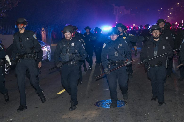 A group of police officers in riot gear with batons walk down a street at night with flashing lights in the background.