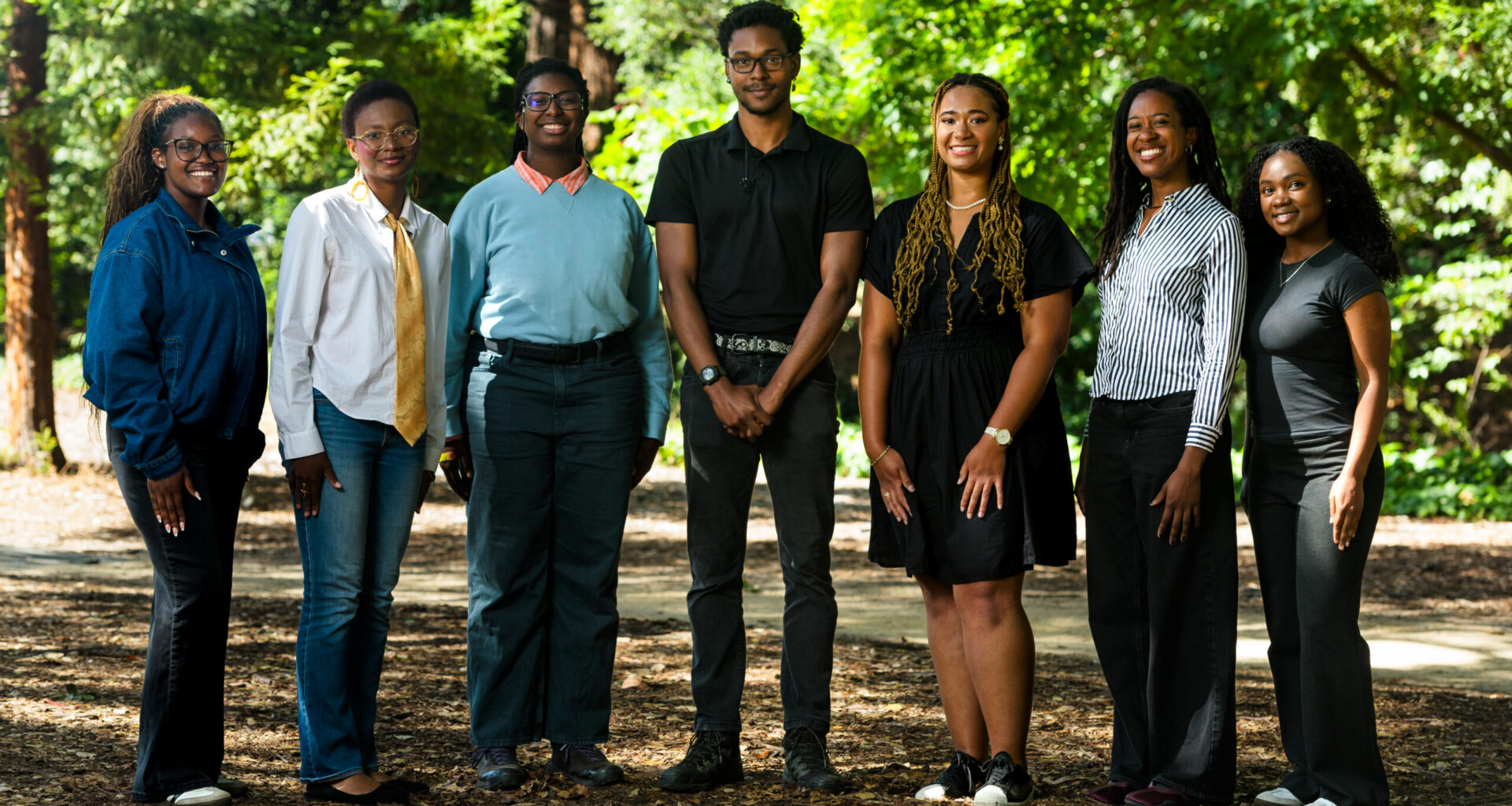 Seven students stand in a row in front of a soft-focus green background outside