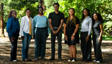 Seven students stand in a row in front of a soft-focus green background outside