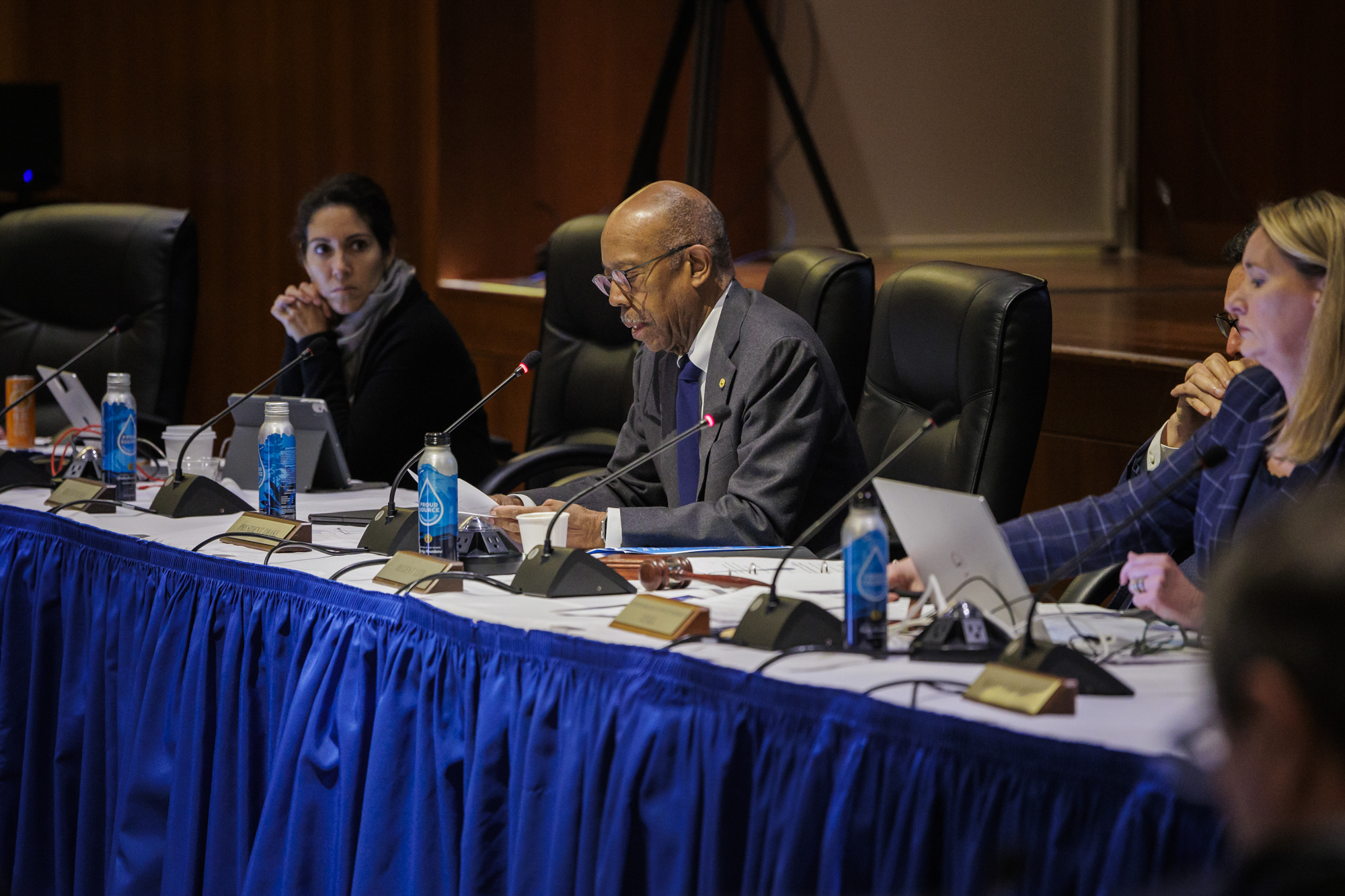 A bald person with glasses speaks into a microphone at a long table.