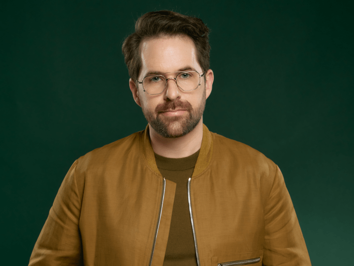 a man poses in a tan jacket in front of a green backgroun
