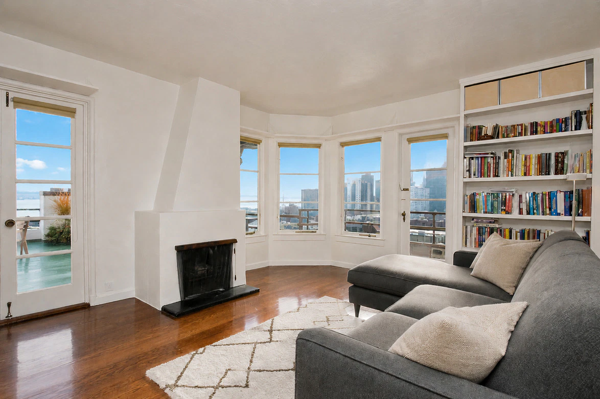 Spacious living room with a fireplace, large windows showcasing a cityscape, and a cozy gray sectional with bookshelves.