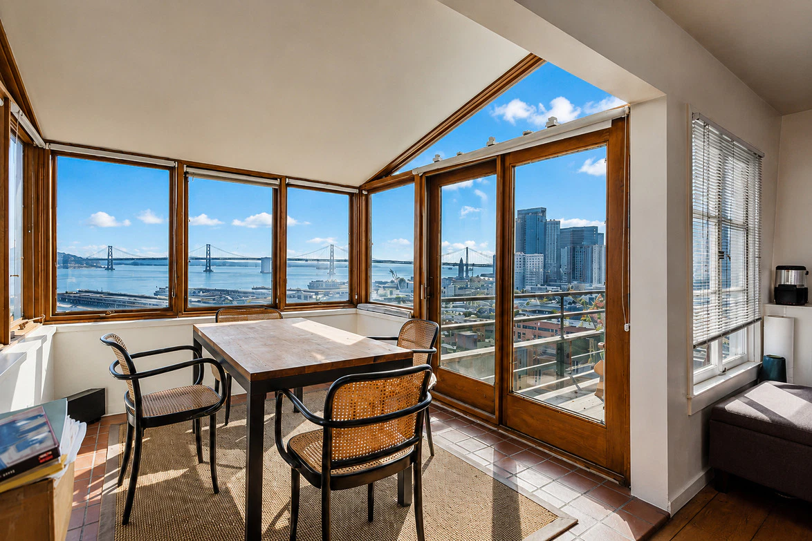 Dining area with wooden table and chairs, large windows overlooking city skyline and a bridge on a sunny day.