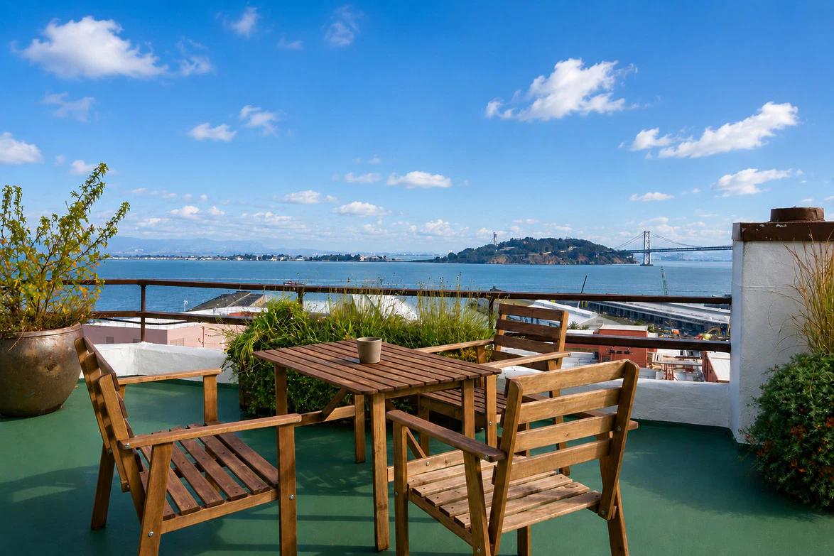 Rooftop patio with wooden chairs and table overlooking the bay and distant bridge on a clear day with scattered clouds.